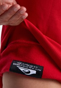 Červená mikina z látky s rebrovanými manžetami a lemom. Čierna nálepka na spodnej časti obsahuje logo a text o výrobe. Ruka je viditeľná, ako ťahá za látku.