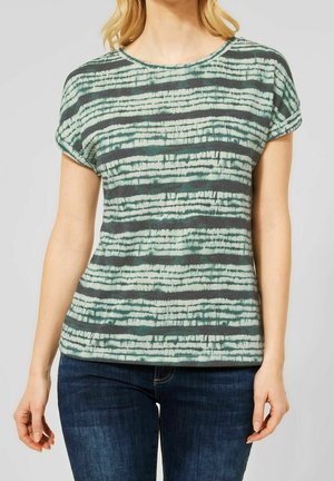 Kvinde iført en kortærmet top med grønne og sorte striber og mørkeblå jeans, stående foran en ensfarvet baggrund.