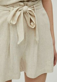 Shorts en lin beige avec une ceinture nouée à la taille, en tissu texturé et coupe légèrement ample, révélant des détails tissés subtils.