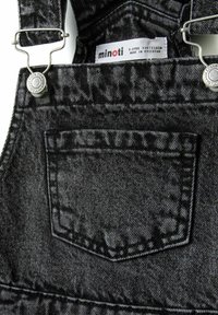 Svarta denimoverall har en framficka, metallklämmor och en etikett som visar storlek och märke. Texturerad tyg med ett något bleknat mönster.