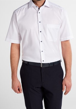 Uomo che indossa una camicia bianca a maniche corte con taschino frontale e pantaloni neri, in piedi contro uno sfondo semplice.