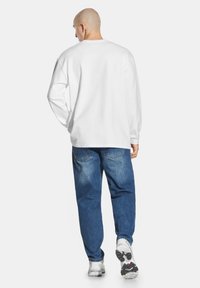 Biała koszula z długim rękawem i okrągłym dekoltem, zestawiona z luźnymi niebieskimi dżinsami i szarymi sneakersami, widok z tyłu.
