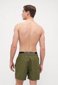 Olíva zöld fürdőnadrág, rugalmas fekete derékkal, amelyen fehér "Calvin Klein" felirat található. Hátoldali zseb részletezve.