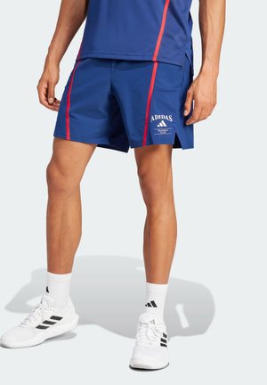 Mężczyzna w niebieskich szortach Adidas z czerwonymi paskami, białych skarpetkach Adidas i białych trampkach stoi na jasnym tle.