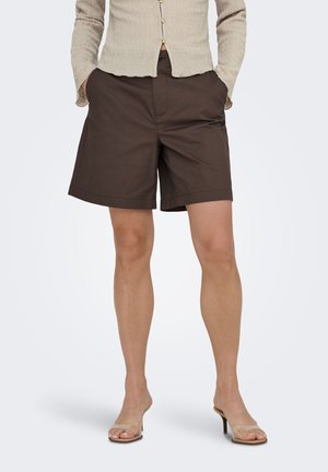 Femme portant un short marron mi-cuisses avec les mains dans les poches, un haut beige texturé boutonné et des sandales à talons avec des brides transparentes.