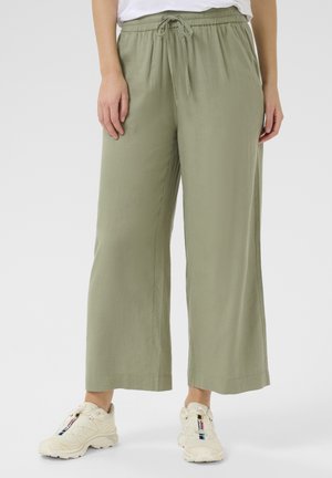 Pantalons larges en vert olive clair en tissu doux avec une taille élastique et un cordon de serrage. Associés à des chaussures de sport beiges.