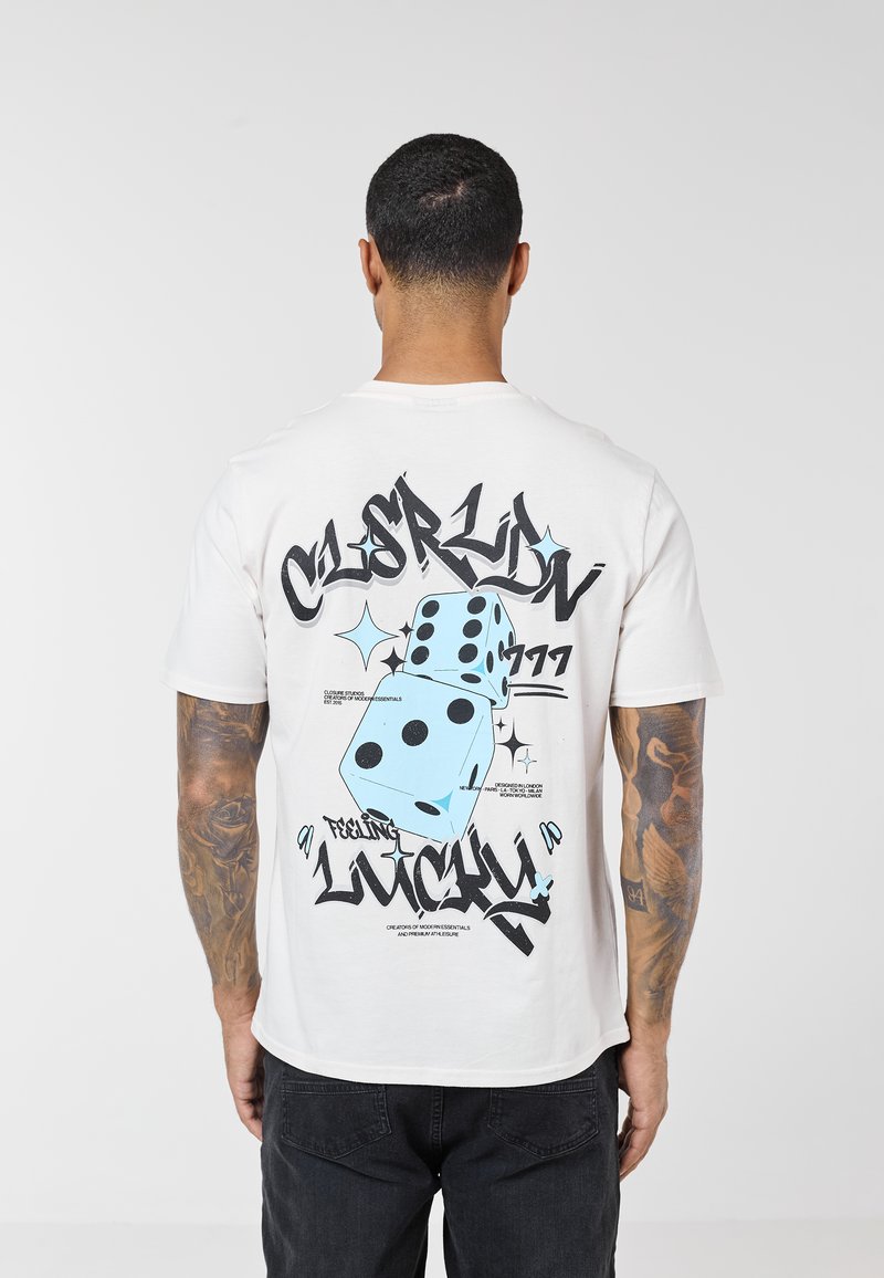 Mand med kort mørkt hår og tatoveringer iført en hvid t-shirt med blå terninger og sort graffiti-tekst, der lyder "Feeling Lucky" på bagsiden.