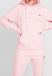 Pink hoodie made of soft fabric with a kangaroo pocket and white drawstrings. Features embroidered logo on the chest. Paired with matching pants.