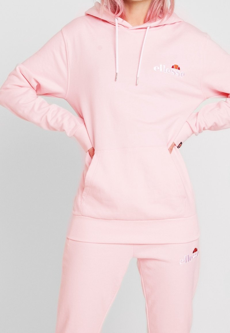 Pink hoodie made of soft fabric with a kangaroo pocket and white drawstrings. Features embroidered logo on the chest. Paired with matching pants.