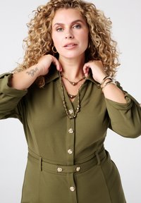 Olijfgroene shirtjurk met een ceintuur in de taille, voorzien van grote gouden knopen en een gladde textuur. Twee gelaagde gouden kettingen met verschillende beads.