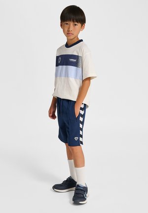 Junge steht mit den Händen in den Taschen, trägt ein weißes und blau gestreiftes T-Shirt, marineblaue Shorts mit Seitenstreifen, weiße Socken und dunkle Turnschuhe.