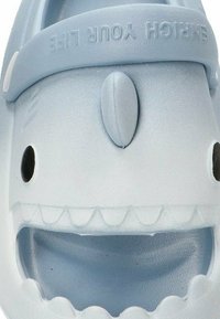 Sandalia de goma azul claro con diseño de cara de tiburón; presenta una parte superior texturizada, forma redondeada, ojos negros y boca bien abierta con acentos de dientes.