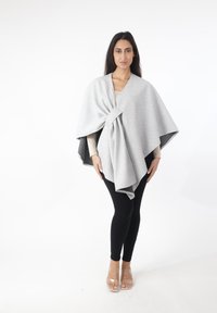 Poncho gris claro con tela texturizada, con un nudo en la parte delantera y detalles en negro a lo largo de los bordes, combinado con leggings negros y sandalias transparentes.