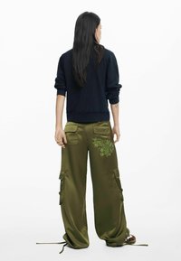 Czarna bluza, zielone satynowe spodnie cargo z kieszeniami oraz ozdobnym haftem kwiatowym. Luźny fason z troczkami przy kostkach.