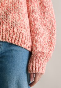 Chunky Strickpullover aus weichem rosa und cremefarbenem Garn, mit lockerer Passform, gerippten Bündchen und einem strukturierten Muster. Kombiniert mit hellblauer Jeans.