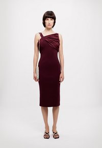 Fioletowa sukienka bodycon z asymetrycznym ramieniem i marszczonym detalem na biuście. Gładka tekstura, długość do kolan, połączona z czarnymi sandałami na cienkich paskach.