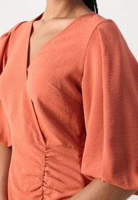 Blazer en terre cuite avec un col en V, des manches bouffantes et des détails froncés à la taille. Tissu lisse de poids moyen avec une texture subtile.