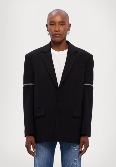 Veste noire en tissu structuré, avec un devant à double bouton, des épaules rembourrées et un accent zippé sur la manche gauche.