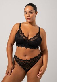 Musta pitsinen bralette, jossa on V-pääntie ja säädettävät hihnat, yhdistettynä vastaavaan pitsiseen thonkiin, jossa on aaltoileva reuna ja monimutkainen kuvio.