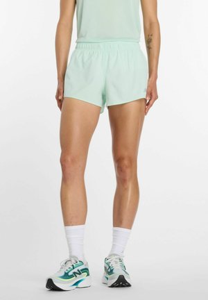 Person iført mintgrønne sportsshorts, matchende ærmeløs top, hvide sokker og turkis-hvide løbesko står mod en ensfarvet baggrund.