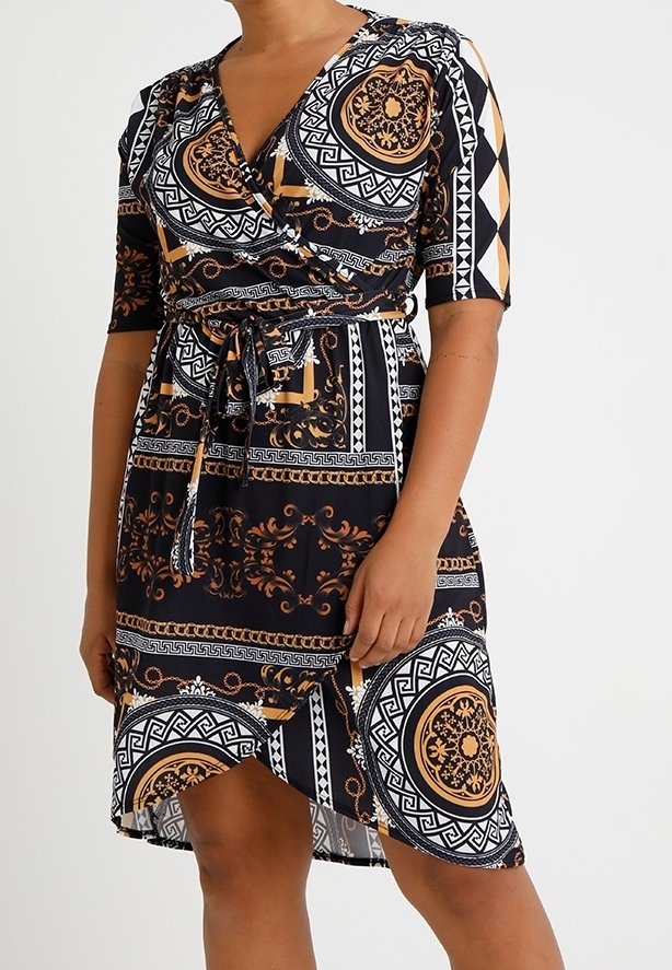 Femme portant une robe portefeuille noire avec des motifs géométriques et floraux complexes dorés et blancs, nouée à la taille, longueur mi-cuisse.