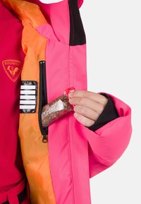 Giacca impermeabile rosa brillante con accenti neri e foderato arancione. Una mano è vista che prende una barretta di cereali da una tasca con zip.