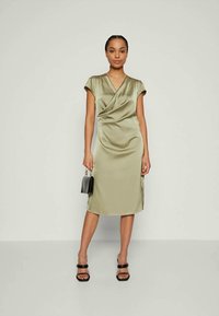 Φόρεμα από σατέν σε ελαιόλαδο πράσινο χρώμα με κοντά μανίκια, σχέδιο τύπου περιτυλίγματος, μέχρι το γόνατο. Συνδυασμένο με μαύρη τσάντα και μαύρες ψηλές σανδάλια.