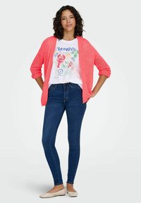 Coral knit cardigan over a white graphic t-shirt featuring lobster design, paired with blue skinny jeans and beige flat shoes.