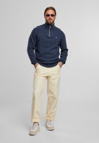 Marineblauer Strickpullover mit hohem Kragen und halbem Reißverschluss, kombiniert mit hellbeigen Hosen und beigen Sneakers.