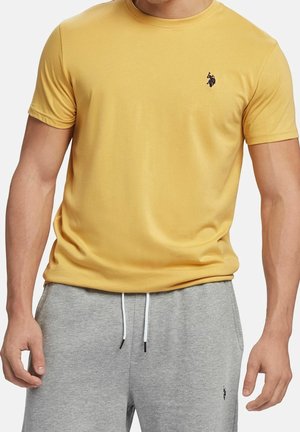 Gelbes kurzärmeliges T-Shirt mit rundem Ausschnitt, aus weichem Material. Verfügt über ein kleines schwarzes Logo auf der linken Brustseite. Wird mit grauen Sweatpants getragen.