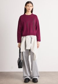 Bordo īsā džemperis ar logotipa druku, kārtots ar gaiši zilu un baltu svītrainu krekla, apvienots ar pelēkiem platām sporta biksēm un melnām kurpēm.