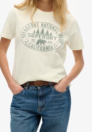 Woman wearing cream t-shirt with "Voyageurs National Park Superdry California" logo and bear graphic, paired with blue jeans and brown belt.