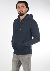 Felpa con zip di colore blu scuro realizzata in tessuto morbido, dotata di cappuccio, tasche frontali e lacci. Design casual con vestibilità slim.