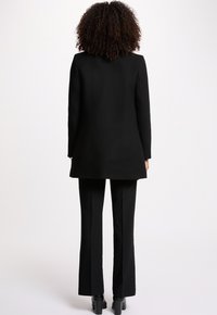 Schwarzer Tailoring-Mantel mit gerader Silhouette, langen Ärmeln und Rückenschlitz, kombiniert mit eng anliegenden schwarzen Hosen und Stiefeletten.