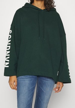 Osoba ubrana w za dużą, ciemnozieloną bluzę z białym nadrukiem na rękawach oraz niebieskie dżinsy, stojąca na białym tle.