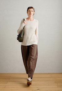 Ljusbeige ribbad tröja, bruna vida byxor och bekväma skor, tillsammans med en grön axelväska. Träddäck och neutral bakgrund.