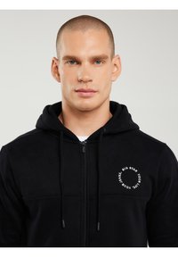 Bărbat cu ochi albaștri, purtând un hanorac negru cu fermoar și text alb circular pe piept, stând în fața unui fundal simplu și deschis la culoare.