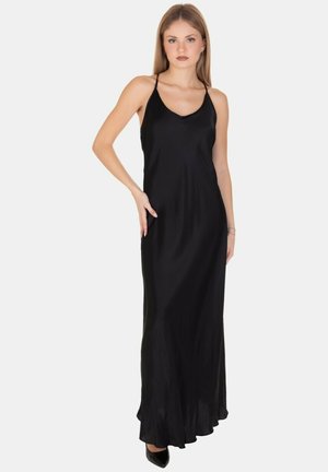 Giovane donna con lunghi capelli, che indossa un maxidress di raso nero senza maniche e tacchi neri, in posa con una mano sulla vita.