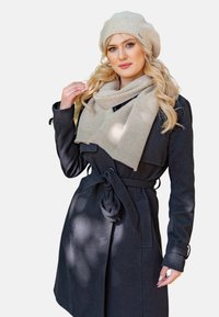 Manteau noir avec une taille attachée, superposé sur une écharpe beige clair. Un béret beige complète la tenue, ajoutant texture et chaleur.