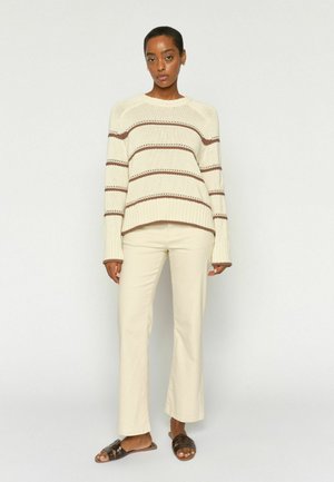 Žena oblečená v krémovom svetri s hnedými pruhmi, krémových nohaviciach a tmavohnedých šľapkách, stojaca pred jednoduchým svetlým pozadím.