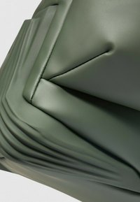 Materiale sintetico verde texturizzato con finitura opaca, caratterizzato da pieghe e cuciture dettagliate che creano una forma strutturata e moderna.