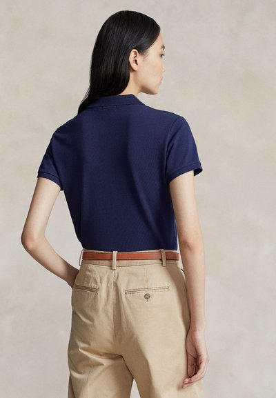 Kvinne med langt svart hår, iført en marineblå kortermet polotrøye stukket inn i beige bukser med brunt belte, sett bakfra.