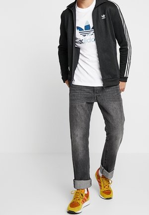 Mies, joka pukeutunut mustaan Adidas-takkiin, valkoiseen Adidas-logolla varustettuun t-paitaan, käännettyihin harmaisiin farkkuihin ja kelta-oransseihin Adidas-tennareihin, seisoo valkoista taustaa vasten.