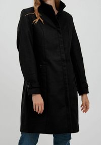 Cappotto di lana nero con colletto alto, chiusura frontale a bottoni, tasche laterali e polsini arrotolati. Texture liscia e design strutturato.