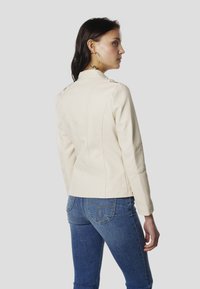 Beige Lederjacke mit strukturierten Nähten, Stehkragen und Schulterklappen. Von hinten mit blauen Denim-Jeans getragen.
