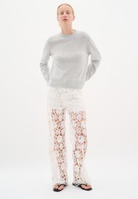 Pulover tricotat gri deschis, cu gât rotund; pantaloni largi albi din dantelă cu modele florale; flip-flops negre pe picioare; fundal neutru.