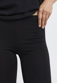 Juodos tamprės su lygiu, tampriu audiniu. Juosmens juosta šiek tiek nuleista, atskleidžiant glaudų prigludimą ir minkštą medžiagą.