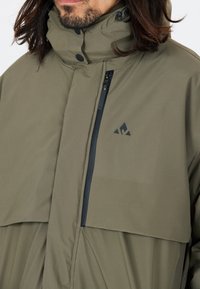 Grön vattentät jacka med hög krage, zip-försedd bröstficka och triangel-logotypdetalj. Matt yta med en minimalistisk design.