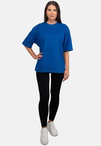 T-shirt in cotone blu con vestibilità rilassata e maniche corte, abbinato a leggings neri e sneakers bianche su uno sfondo neutro.