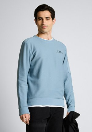 Man draagt een lichtblauwe trui met lange mouwen en een zwarte broek, houdt een opgevouwen zwarte jas vast, staat tegen een effen lichte achtergrond.